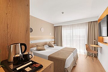 A bright, modern hotel room with two beds, a coffee station, a desk, a wall-mounted TV, and a large window with sheer curtains.