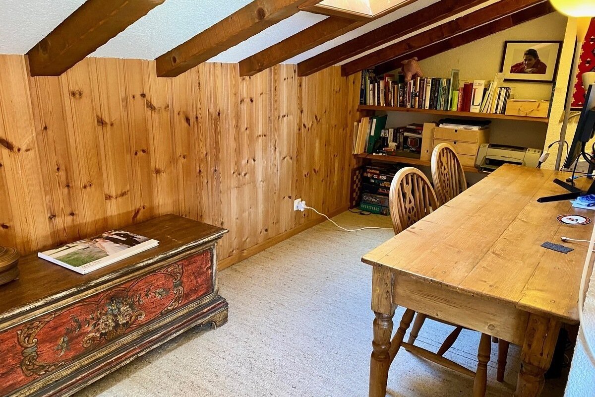 A cozy, wood-paneled attic office with exposed beams, a large wooden desk, bookshelves, a printer, and a decorative chest on a carpeted floor.