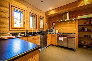 A cozy, rustic kitchen features warm wooden walls and cabinets, black countertops, stainless steel appliances, and bright windows, creating an inviting atmosphere.