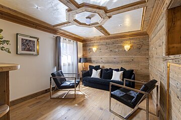 A cozy living room features rustic wood walls, ceiling, and floor. A dark blue sofa, modern black chairs, and a deer artwork complete the space.