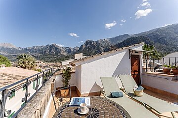 Sunny rooftop terrace with stunning mountain views, loungers, a mosaic table, and a coffee cup, under a clear blue sky.
