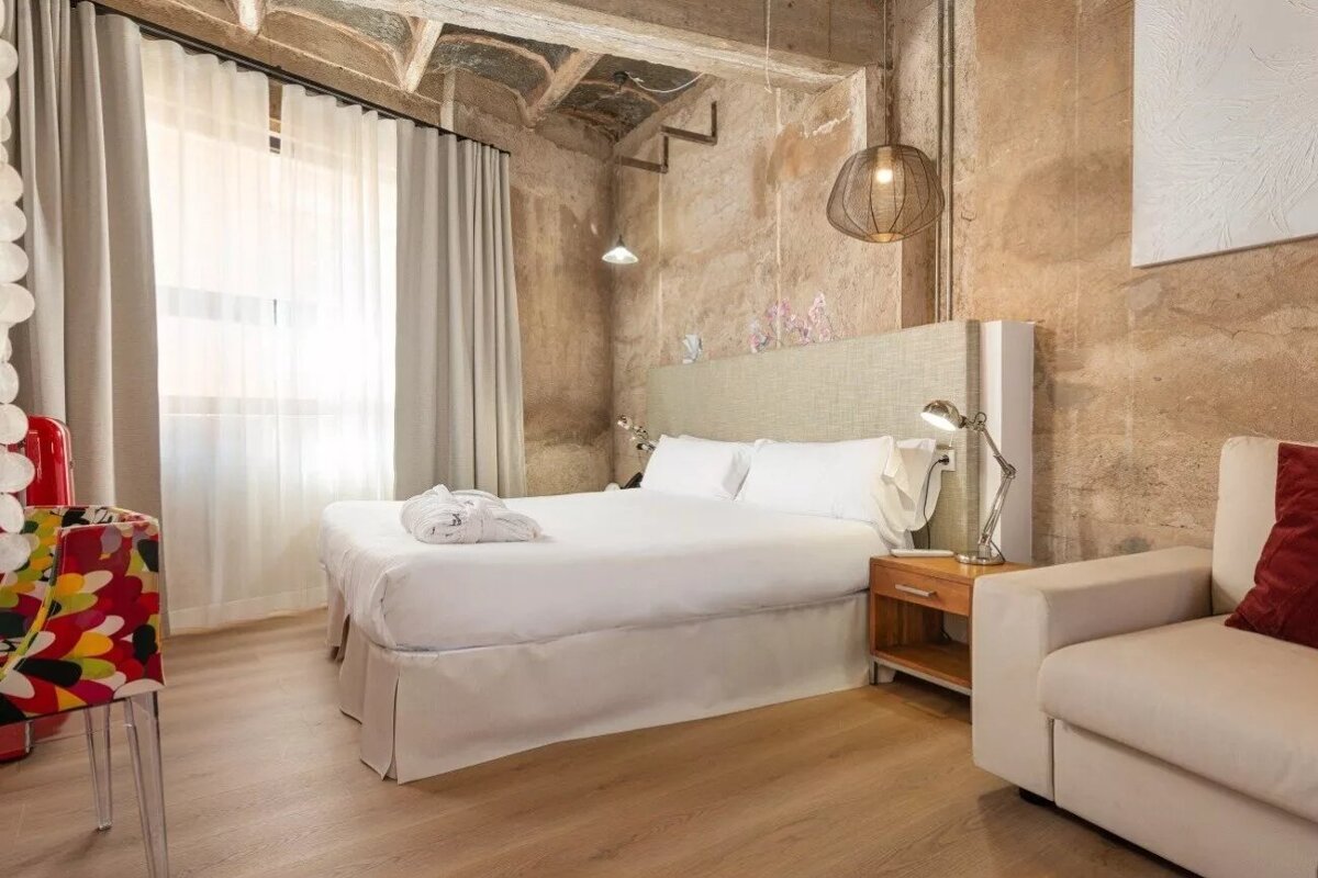 Modern hotel room with a white bed & sofa, rustic concrete walls, wooden floor, & window. A colorful accent chair adds a vibrant touch.