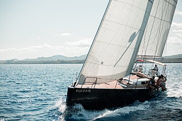 A sailboat with full sails and people aboard glides on sparkling blue water past a sunny coastal landscape with distant mountains.