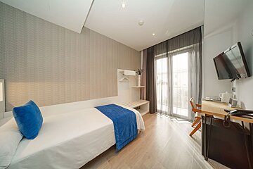 A bright, modern hotel room with a single bed, blue accents, a desk with a TV, and a balcony with sheer curtains.