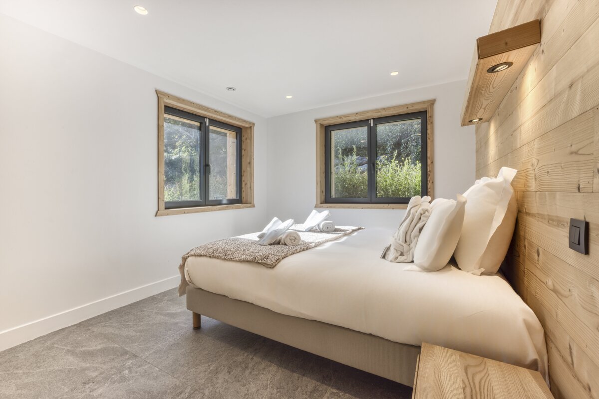 A modern bedroom features a double bed with white bedding, two windows overlooking greenery, and a wooden accent wall with integrated lighting. Grey tiled floor completes the clean look.