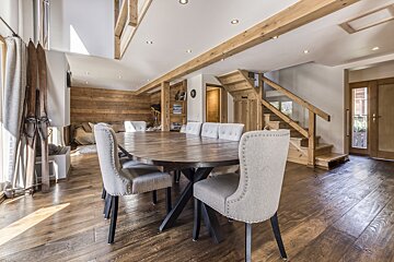 A bright, rustic chalet interior with a large wooden dining table, upholstered chairs, wood-paneled walls, exposed beams, and a wooden staircase.