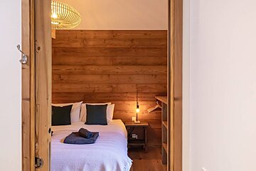 A cozy, rustic bedroom with warm wooden walls, a bed with dark pillows, and a bedside lamp, seen through a wooden doorway.