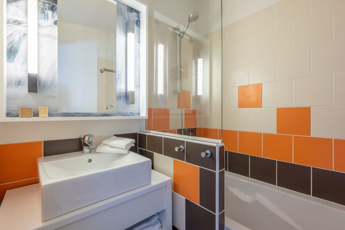 A modern bathroom with a white vessel sink, a large lighted mirror, and a tiled shower-tub area with orange and brown accents.