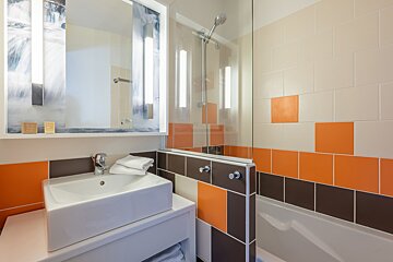 A modern bathroom with a white vessel sink, a large lighted mirror, and a tiled shower-tub area with orange and brown accents.