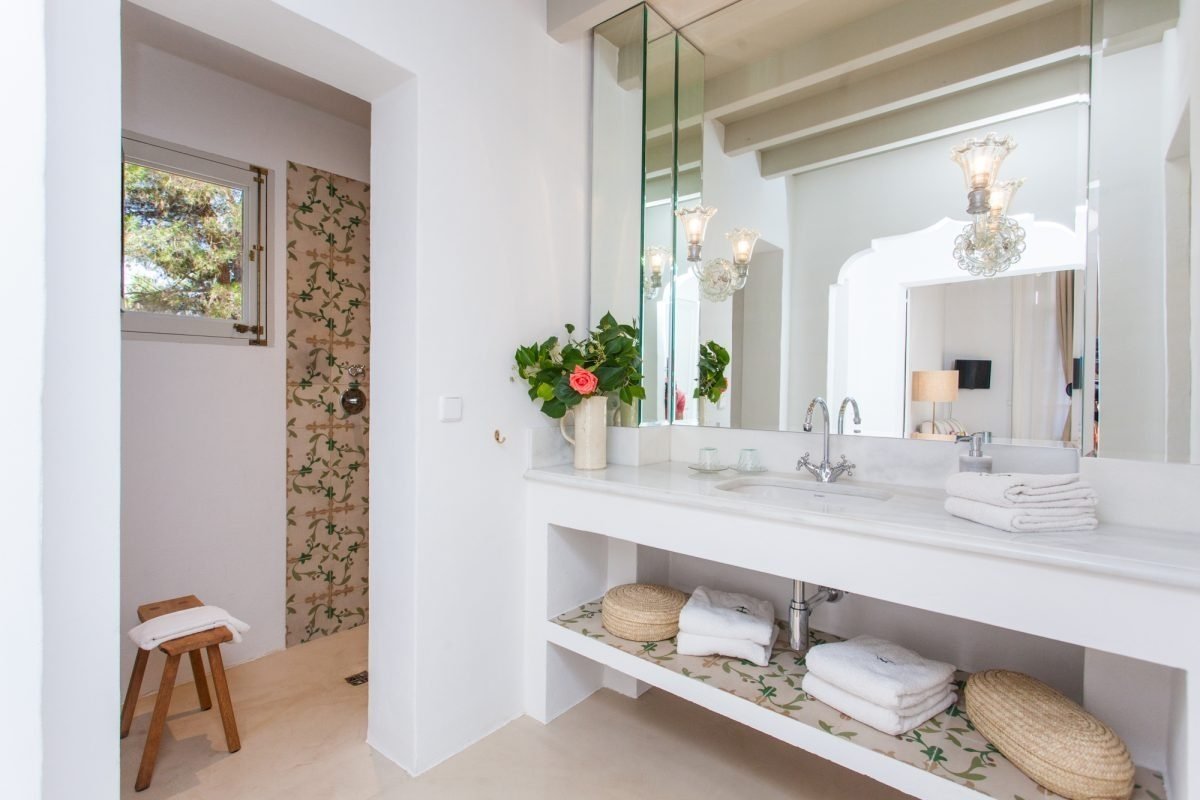 A bright white bathroom with a double sink vanity, large mirror, and a shower area featuring decorative green and beige tiles. A rose bouquet adds color.