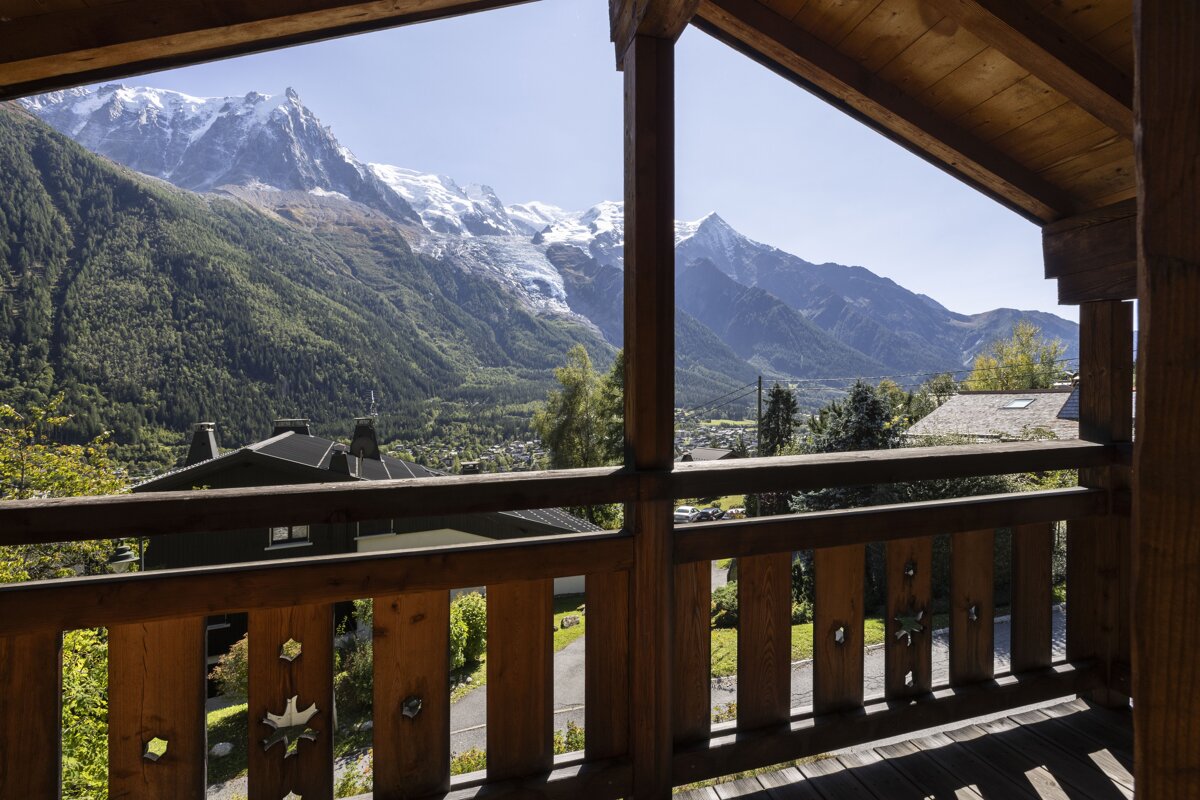 Mountain view from a wooden balcony, featuring snow-capped peaks, a glacier, a green valley, and a village nestled below.