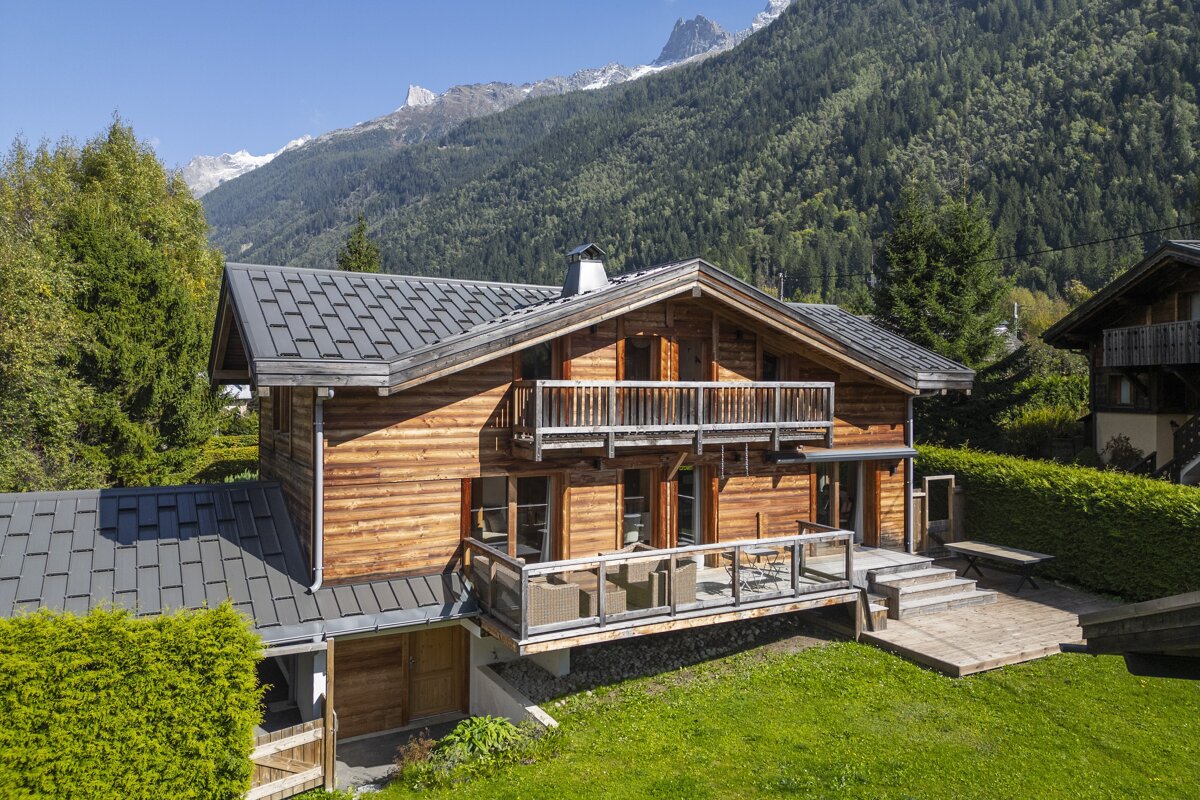 A charming wooden chalet with balconies, set amidst green lawns and towering, forested mountains with snowy peaks under a clear blue sky.