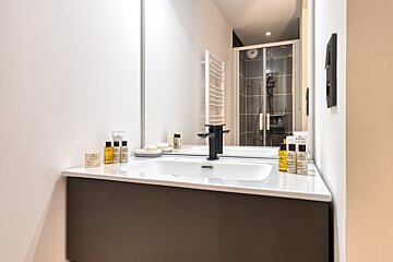 A sleek bathroom features a white vanity, black faucet, and Nuxe toiletries. The mirror reflects a shower and white towel warmer.