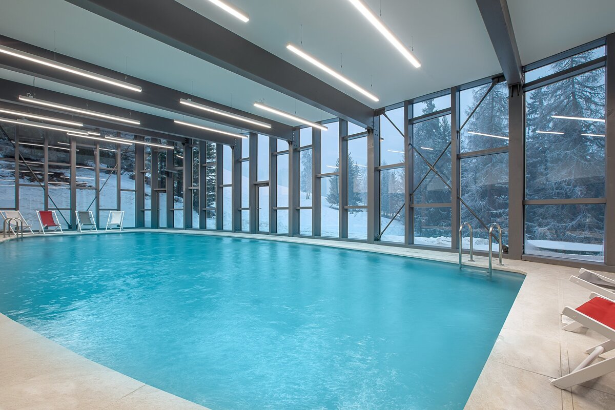 A modern indoor swimming pool with large windows overlooks a snowy, forested landscape. Bright linear lights illuminate the serene blue water and modern architecture.