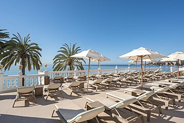 A sunny oceanfront terrace with numerous lounge chairs, white umbrellas, and palm trees, overlooking a clear blue sea.