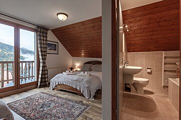 A cozy attic bedroom with a bed, patterned rug, and balcony offering misty mountain views. An en-suite bathroom with a toilet and sink is visible to the right.