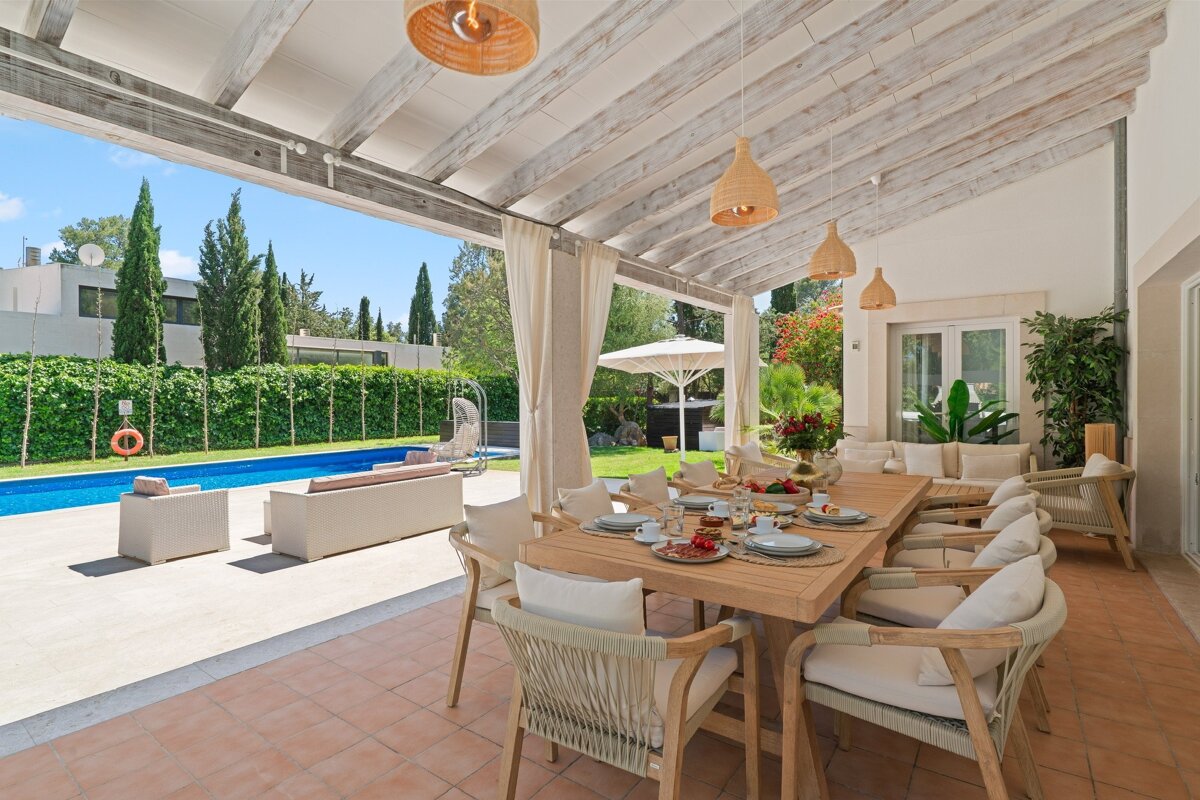 Bright outdoor dining patio with a large table set for a meal, overlooking a blue swimming pool, lush garden, and modern house.