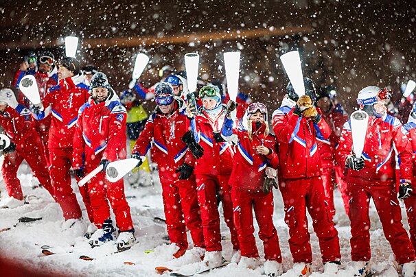 Skiers in red suits stand in heavy snowfall at night, holding illuminated white batons. Snow covers the ground and falls thickly around them.