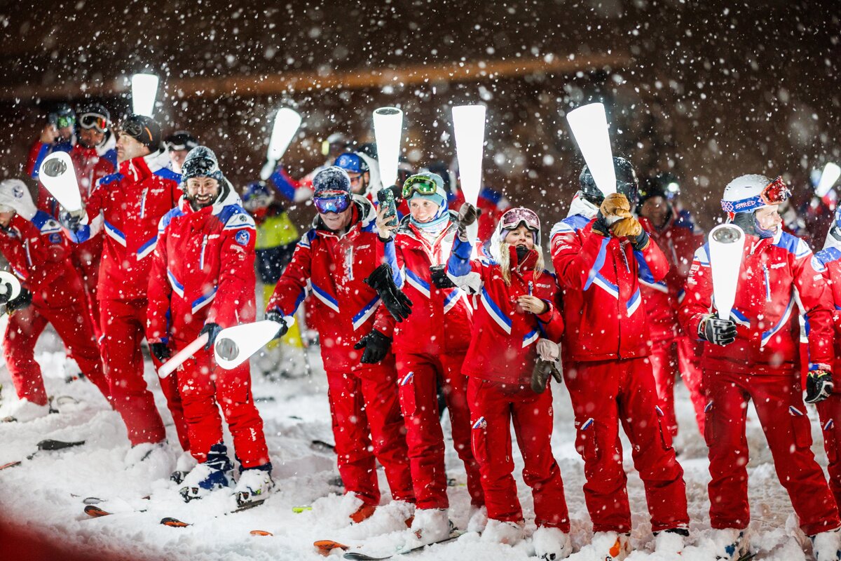 Skiers in red suits stand in heavy snowfall at night, holding illuminated white batons. Snow covers the ground and falls thickly around them.