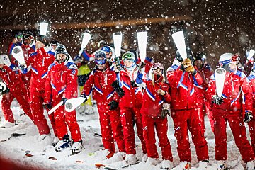 Skiers in red uniforms hold glowing white light sticks while standing in heavy snowfall at night, appearing excited amidst the flakes.