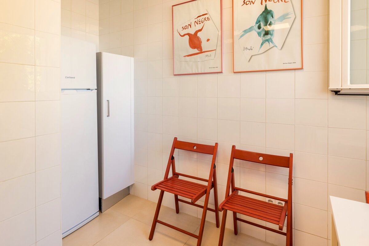 A white tiled room with a fridge, cabinet, two red folding chairs, and two framed graphic posters hanging on the wall.