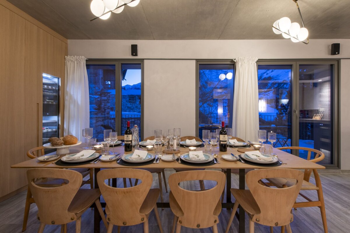 A modern dining room with a long, set wooden table and contemporary lighting. Large windows overlook a dark, wintery outdoor landscape.