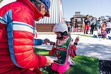 A ski instructor awards a badge to a young girl in pink ski gear, kneeling on artificial turf in a snowy mountain resort.