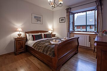 A cozy bedroom with a classic wooden sleigh bed, furry throw, and illuminated lamps. An open window provides a view of a charming stone building outside.