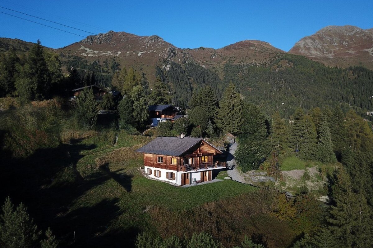 A rustic wooden chalet on a green, forested mountain slope with other homes and peaks under a clear blue sky.