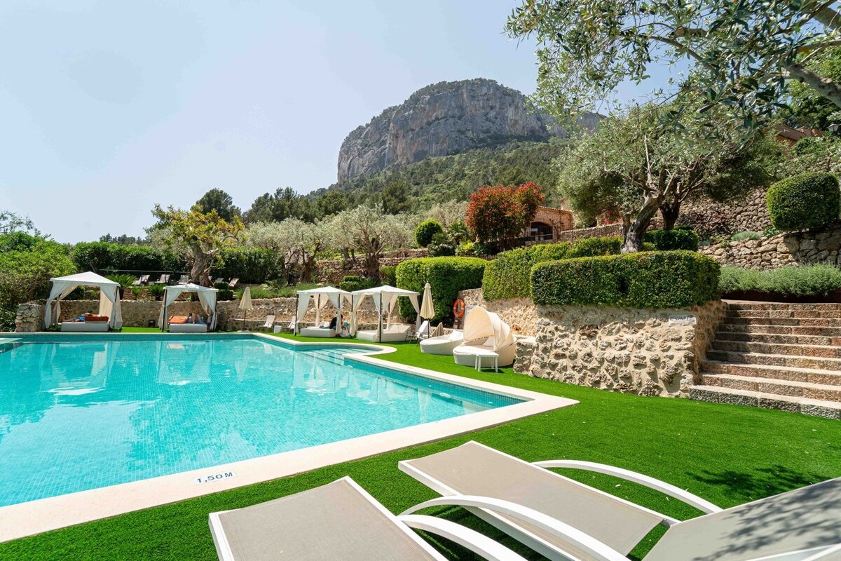 A luxurious outdoor pool area with cabanas, lounge chairs, lush greenery, and a majestic mountain backdrop under a clear sky.