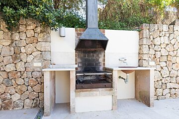 An outdoor BBQ area with stone walls, a built-in brick grill with metal hood, and a sink with a rustic basin and faucet.