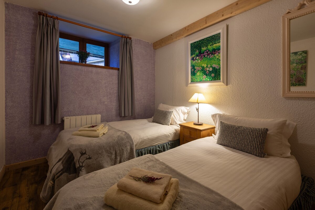 A cozy twin bedroom features two single beds, a purple textured wall, a window with grey curtains, and artwork. A bedside lamp illuminates the space.