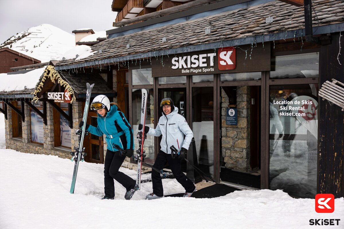 Two people carrying skis in front of a ski set store