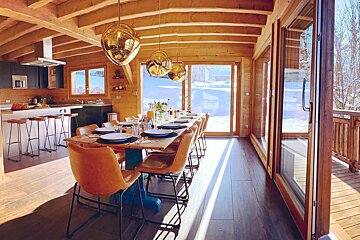 A bright, open-plan wooden chalet interior features a modern kitchen, a long dining table set for guests, and large windows overlooking a snowy mountain landscape.