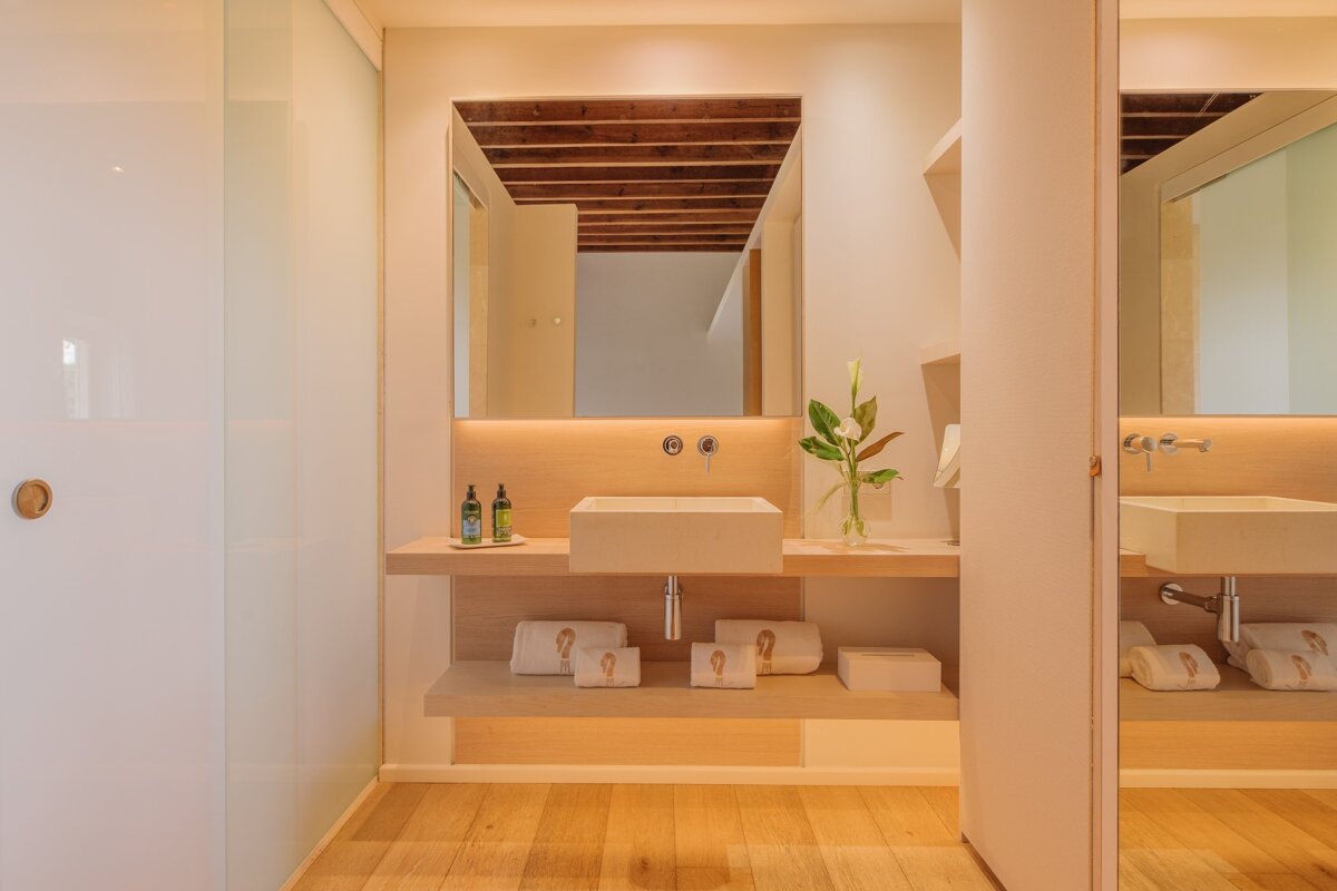 A minimalist bathroom featuring a light wood vanity, square sink, and backlit mirror reflecting a wooden beamed ceiling. White towels & toiletries complete the clean look.