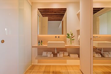 A minimalist bathroom featuring a light wood vanity, square sink, and backlit mirror reflecting a wooden beamed ceiling. White towels & toiletries complete the clean look.