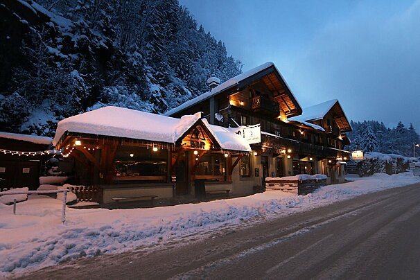 A cozy, snow-covered chalet with warm glowing lights at dusk. A snowy road runs past, flanked by dark, forested mountains under a dusky sky.