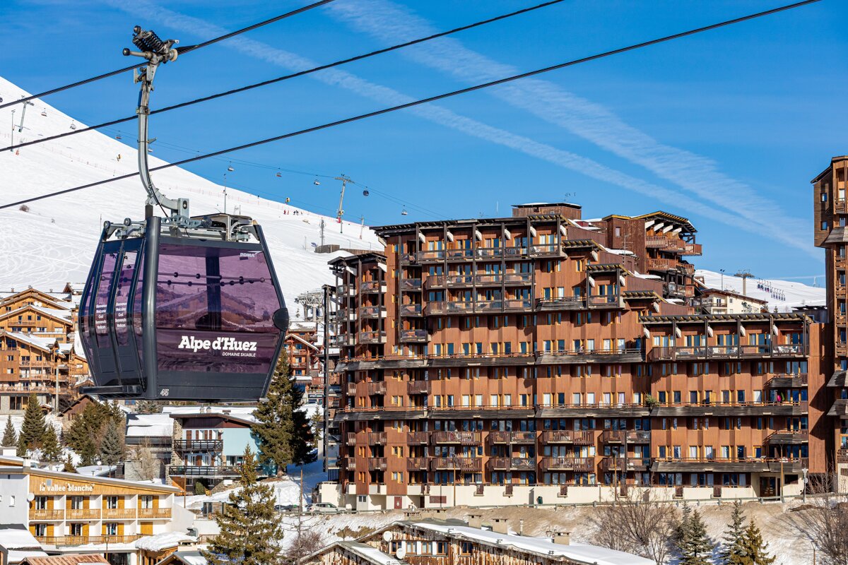 A purple Alpe d'Huez cable car hanging over a snowy ski resort with large wooden buildings under a clear blue sky.