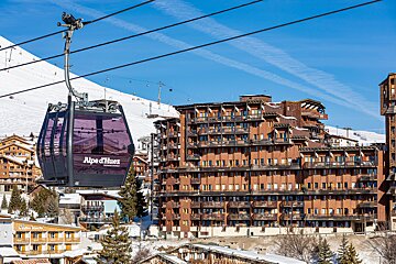 A purple Alpe d'Huez cable car hanging over a snowy ski resort with large wooden buildings under a clear blue sky.