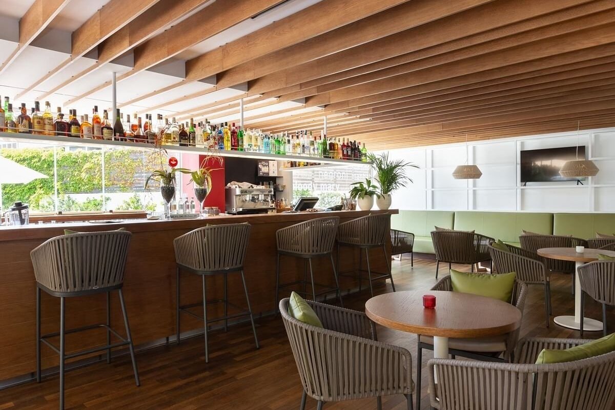 Modern bar and lounge featuring a wooden bar, liquor bottles, chic seating with green cushions, and a striking slatted wooden ceiling.
