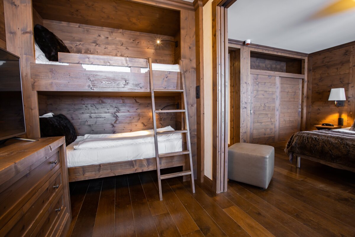 Cozy, wood-paneled room with integrated bunk beds, a TV, and a ladder. An adjacent bedroom with a double bed and warm lamp is visible through a doorway.