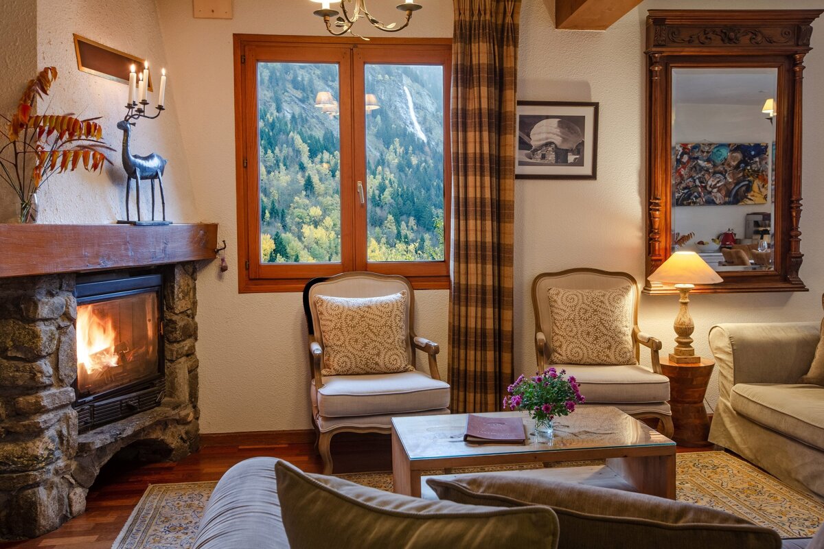 A cozy living room features a roaring fireplace, classic armchairs, and a large window showcasing a vibrant autumn forest. Warm and inviting ambiance.