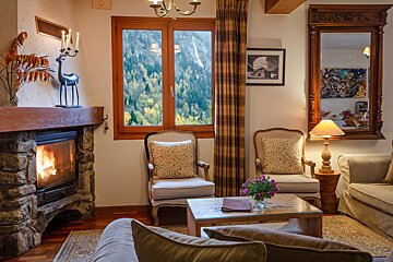 A cozy living room features a roaring fireplace, classic armchairs, and a large window showcasing a vibrant autumn forest. Warm and inviting ambiance.