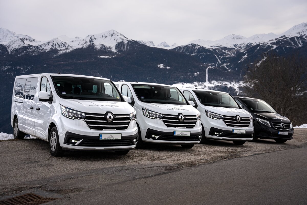 A row of white vans are parked in front of snowy mountains