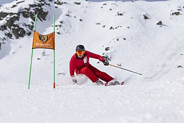 A red-suited skier carves a turn on a snowy mountain, kicking up powder next to an orange ski race gate.