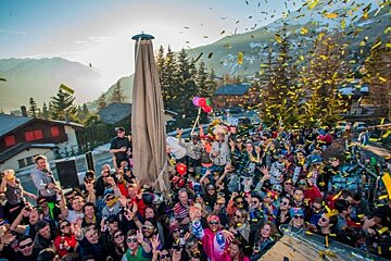 A huge, festive crowd celebrates outdoors with falling yellow confetti against a snowy mountain and village backdrop.