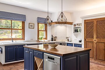 A rustic-modern kitchen featuring navy blue cabinets, a wooden island, terracotta floor, and sunlit windows. Geometric pendant lights add a chic touch.
