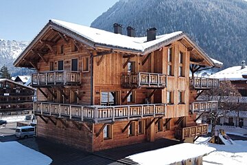 A large, multi-story wooden chalet with snow-covered roofs and numerous balconies, set amidst a snowy mountain landscape.