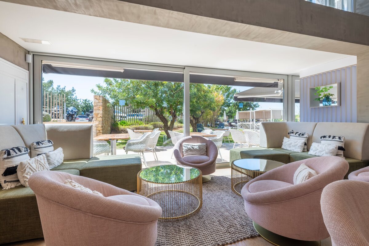 A bright indoor lounge with pink and green seating, glass tables, overlooking a sunny outdoor patio through large windows.