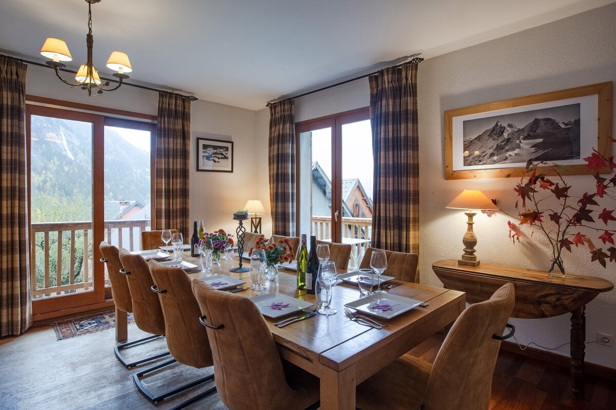 A charming dining room with a large wooden table set for dinner, offering a stunning mountain view from the balcony. Plaid curtains and warm lighting create a cozy ambiance.